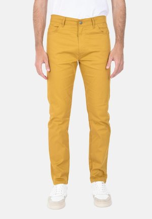 Uomo che indossa pantaloni slim-fit giallo senape e sneakers bianche, in piedi davanti a uno sfondo chiaro e uniforme.