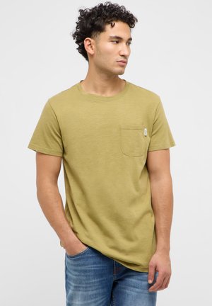 Man with curly hair wearing an olive green crew-neck t-shirt with a chest pocket and blue jeans, looking to his right.