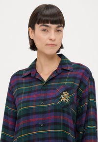 Zelena in vijolična kockasta srajca na gumbe s ovratnikom, ki ima na levem žepu zlat vezene logotip in črne gumbe.