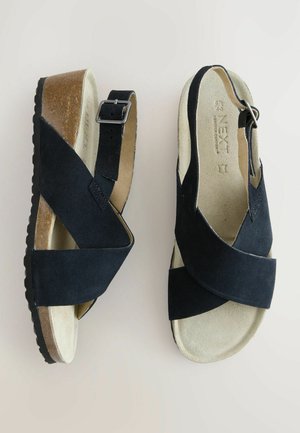 Crne nubuk sandale s prekriženim trakama, podesivom kopčom i laganim podmetanjem od pluta. Mekana unutarnja postava osigurava udobnost.