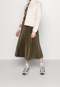 Osoba ubrana w beżową kurtkę puchową, kwiatową bluzkę, oliwkową plisowaną midi spódnicę, różowe skarpetki i białe z zielonymi sneakersami, stojąca na gładkiej podłodze.