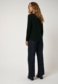 Dunkelgrüner Pullover kombiniert mit lässigen, lockeren dunklen Denimhosen. Der Pullover hat lange Ärmel und eine glatte Textur; die Hosen weisen sichtbare Nähte auf.