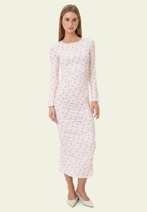 Kvinde står iført en lyserød midi-kjole med lange ærmer og små blomsterprint samt beige spidse hæle mod en ensfarvet baggrund.