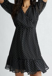 Mustat pilkulliset mekko, joka on valmistettu kevyestä kankaasta. Mekossa on kietaisumalli, levenevät hihat ja röyhelöhelma. Pilkut ovat valkoisia.