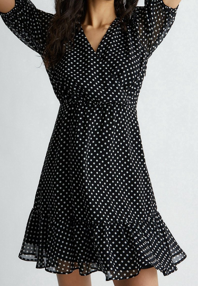 Mustat pilkulliset mekko, joka on valmistettu kevyestä kankaasta. Mekossa on kietaisumalli, levenevät hihat ja röyhelöhelma. Pilkut ovat valkoisia.