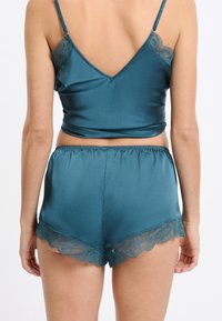 Satynowa kamizelka w kolorze morskim z cienkimi ramiączkami i koronkowymi zdobieniami; dopasowane szorty z falistym brzegiem i koronkowym wykończeniem. Gładka faktura, dopasowany krój.