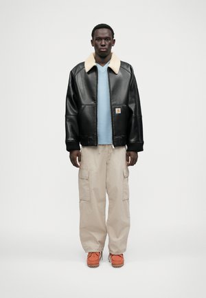 Czarna skórzana kurtka bombowiec z kremowym kołnierzem z owczej wełny, noszona na jasnoniebieskiej koszuli, w zestawie z beżowymi spodniami cargo i pomarańczowymi butami.