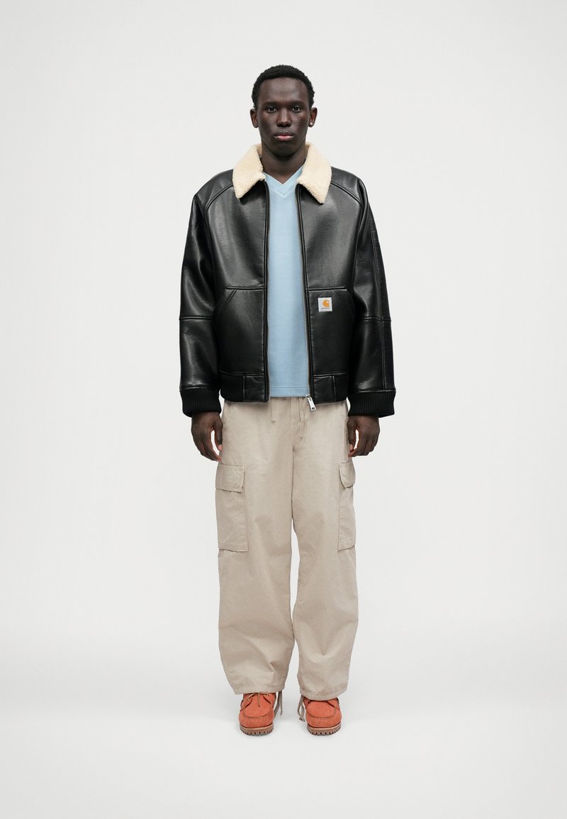 Czarna skórzana kurtka bombowiec z kremowym kołnierzem z owczej wełny, noszona na jasnoniebieskiej koszuli, w zestawie z beżowymi spodniami cargo i pomarańczowymi butami.