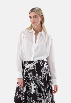 Blonde Frau trägt transparente weiße Bluse mit Knopfleiste und hoch taillierten schwarzen Rock mit großem weißem Blumenmuster, posiert vor weißem Hintergrund.