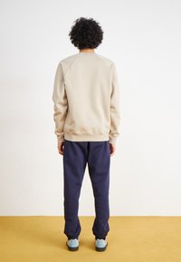 Beige Sweatshirt mit Raglanärmeln, kombiniert mit navyblauen Sweatpants, die elastische Bündchen haben, und grauen Sneakers, von hinten betrachtet.