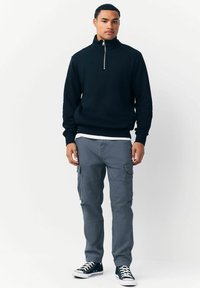 Námořnická mikina na zip s vysokým límcem, spárovaná se šedými cargo kalhotami. Kalhoty mají boční kapsy a volný střih. Černé tenisky.