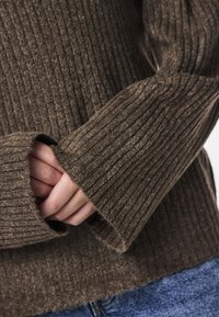 Mains croisées à l'intérieur de longues manches côtelées d'un pull marron, portant un jean bleu, vue en gros plan du haut du torse et des bras.
