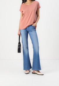 Różowa bluzka z krótkim rękawem, niebieskie dżinsy z rozkloszowanymi nogawkami, beżowe baleriny i czarna torebka. Gładki materiał z podwiniętymi rękawami i luźnym krojem.