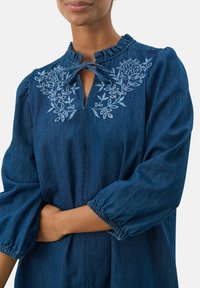 Denim blúz fodros gallérral, kulcslyuk előrésszel megkötő részletekkel, valamint világoskék virágos hímzéssel. Hosszú ujjú, gumírozott mandzsettával.