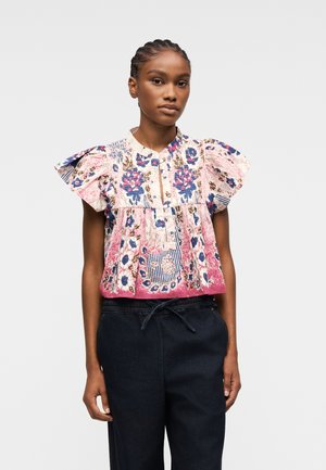 Frau trägt eine kurzärmelige Blumenbluse mit Rüschen an den Schultern und dunkle Hose mit Kordelzug, steht vor einem schlichten Hintergrund.