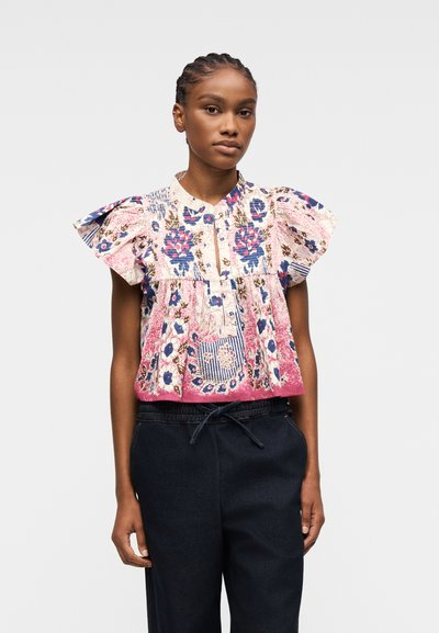 Woman wearing a short-sleeve floral blouse with ruffled shoulders and dark drawstring pants, standing against a plain background.