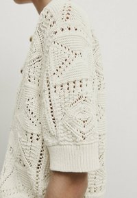 Cremfarbener, gestrickter Pullover mit einem strukturierten Muster aus Rautenformen, kurzen Ärmeln und gerippten Bündchen.