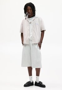 Camicetta a maniche corte a righe beige e bianche, abbinata a pantaloni corti grigi al ginocchio e scarpe nere. Presenta una vestibilità casual e dettagli minimi.