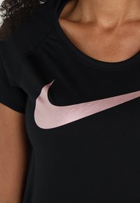 Camiseta atlética negra de manga corta con un brillante logo de Nike en rosa dorado en el pecho, que muestra una tela suave y ligera.