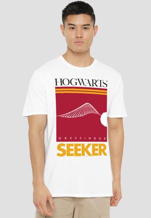 Young man wearing a white t-shirt with red and gold Hogwarts Gryffindor Seeker design and beige shorts, standing against a plain background.