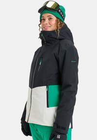 Schwarze und weiße Skijacke mit grünen Akzenten, ausgestattet mit einem hohen Kragen, Reißverschlüssen und Logodetail am Ärmel. Glattes, wetterfestes Material.