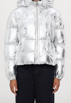 Osoba ubrana w błyszczącą srebrną puchową kurtkę z kapturem, suwakiem z przodu i elastycznymi mankietami, stojąca na jednolitym tle, ubrana w ciemne spodnie.