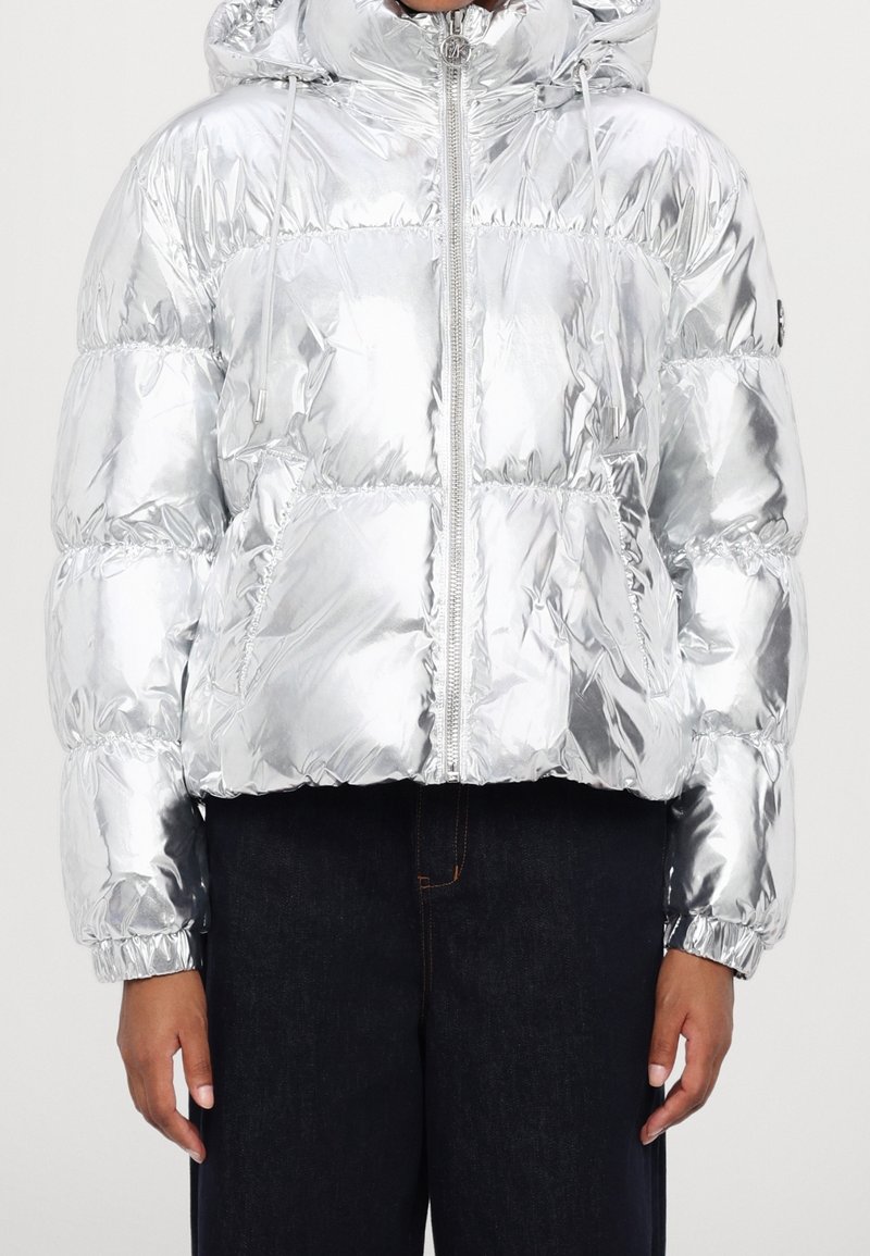 Personne portant une veste matelassée argentée brillante avec une capuche, une fermeture éclair à l'avant et des poignets élastiques, sur un pantalon foncé, debout devant un fond uni.
