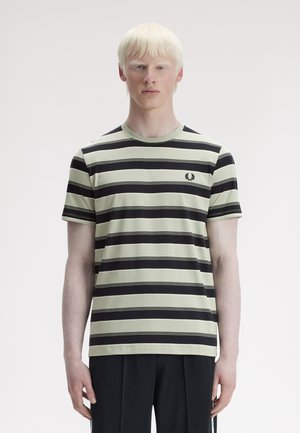 Personne aux cheveux blonds très clairs et courts, portant un t-shirt à rayures horizontales noires et blanches et un pantalon noir, devant un fond uni.