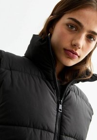 Chaqueta de plumas negra con un cuello alto, cierre de cremallera y textura acolchada. Presenta un material suave y brillante, sin patrones visibles.