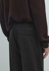 Pantaloni grigio scuro su misura con righe verticali, fronte piatto e una tasca sul retro, abbinati a un maglione marrone con texture.