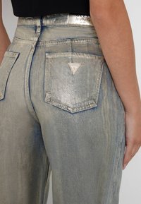 Wycierane niebieskie dżinsy z metalicznym połyskiem, z trójkątną łatą z logo na tylnej kieszeni oraz wysokim stanem.