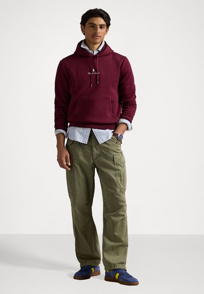 Um hoodie bordeaux com um bolso, sobre uma camisa riscada azul clara, combinado com calças cargo verde-oliva e sapatilhas desportivas azuis com detalhes amarelos.