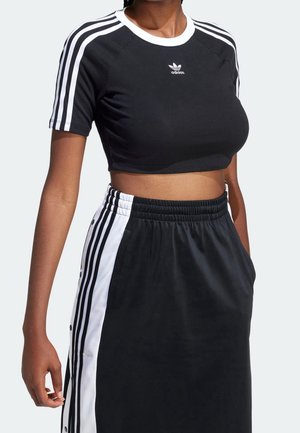 Kobieta ubrana w czarny krótki T-shirt Adidas z białymi paskami na rękawach oraz dopasowaną czarną spódnicę z białymi bocznymi panelami i paskami.