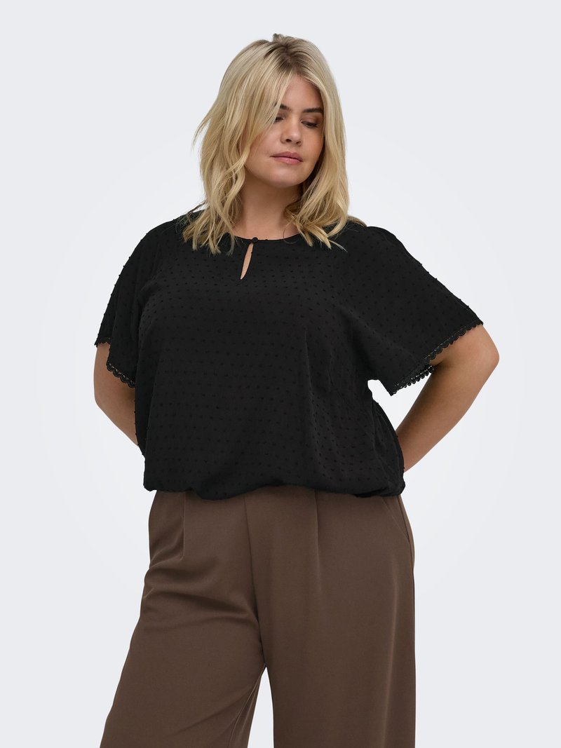 Femme aux cheveux blonds portant un chemisier noir à manches courtes avec de petits pois et un pantalon marron à jambes larges, debout les mains dans le dos.