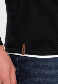 Black knitted sweater with a textured finish, featuring a brown leather label with "ombre" at the hem, layered over a white undershirt.
