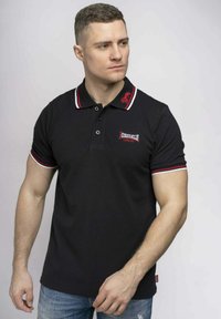 Fekete polo ing piros és fehér csíkos gallérral és mandzsettával, mellrészen logó található; pamutból készült, testhezálló dizájnnal.