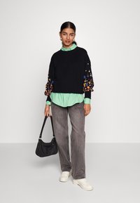 Μαύρος πουλόβερ με πολύχρωμη floral κεντητική στα μανίκια, επικαλυμμένο με ένα ανοιχτό πράσινο πουκάμισο με κουμπιά, σε συνδυασμό με γκρι τζιν και λευκά αθλητικά παπούτσια.