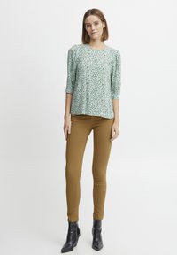 Bloemmotief, groene blouse met driekwartmouwen, gecombineerd met aansluitende, mosterdkleurige broeken. Zwarte enkellaarsjes met een sleehak maken de look compleet.