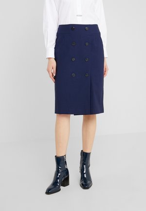 Vrouw draagt een knielange marineblauwe dubbelrijige rok, een wit overhemd met lange mouwen en marineblauwe lakleren enkellaarzen.