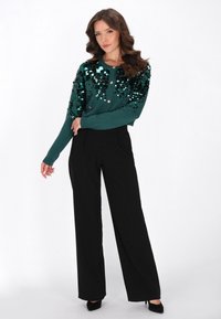 O femeie cu păr ondulat de culoare castanie poartă un cardigan verde împodobit cu paiete, pantaloni negri cu picior larg și sandale negre cu toc, pozând pe un fundal alb.