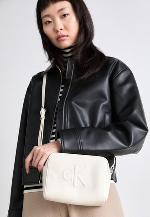 Jeune femme portant une veste en cuir noire et une chemise rayée tient un sac à épaule crème Calvin Klein.