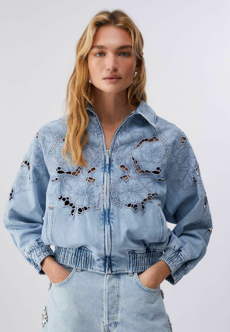 Fiatal nő hosszú szőke hajjal, világoskék farmerdzsekit visel, amely virágmintás kivágott hímzéssel díszített, zsebeibe tett kézzel áll egyszínű háttér előtt.