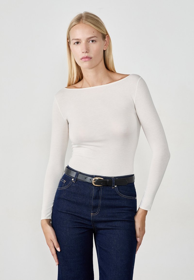 Langarm, off-white top van zacht materiaal, gecombineerd met donkerblauwe jeans en een zwarte riem met een ronde gesp. Soepele textuur en een getailleerd ontwerp.