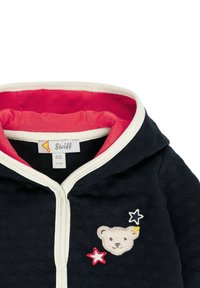 Sweat à capuche marine avec une doublure rose, des bordures blanches et un ours brodé avec des étoiles colorées. L'étiquette indique la taille 62 (2-3 mois). Texture de tissu douce.
