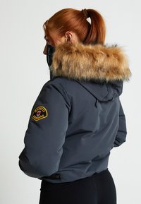 Pūkains tumši pelēks mētelis ar ādas oderi, piedāvājot uzšuvumu uz piedurknes un regulējamu aprīkojumu jostas daļā. Gluda tekstūra.