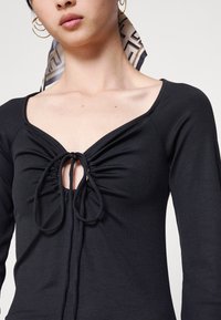 Top preto de mangas longas com decote em V profundo e tecido franzido na frente, apresentando um fecho com cordão e uma textura suave.