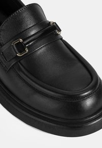 Schwarze Lederloafer mit abgerundetem Zehen, erhöhten Nähten, metallischen Schnallenakzenten und einer klobigen Sohle. Glatte Textur mit polierter Oberfläche.