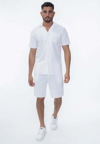Chemise blanche à manches courtes avec un bouton et une poche poitrine, associée à un short blanc, portée avec des baskets blanches, présentée sur un fond neutre.