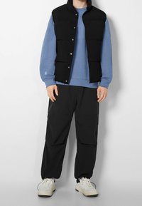 Černá péřová vesta přes světle modrou mikinu, v kombinaci s černými zúženými kalhotami a bílými teniskami. Měkké textury a ležérní styl.