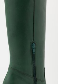 Botte haute en cuir vert avec une fermeture éclair latérale, une texture lisse et des détails cousus le long des coutures pour une apparence sur mesure.